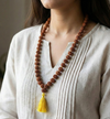 Person wearing a long brown beaded necklace with a yellow tassel on a neutral background