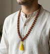 Man wearing a long brown beaded necklace with a yellow tassel on a neutral background