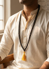 Man wearing a beige kurta with a long black beaded necklace and yellow tassel.