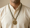 Man wearing a black beaded necklace with a gold pendant on a beige shirt.