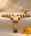 Necklace with rudraksha beads and a gold pendant on a textured surface with flowers and a lamp in the background.