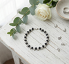 Black beaded bracelet on a white wooden surface with a white rose and green leaves.