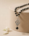 Black beaded necklace with a silver pendant on a beige surface with a white flower.